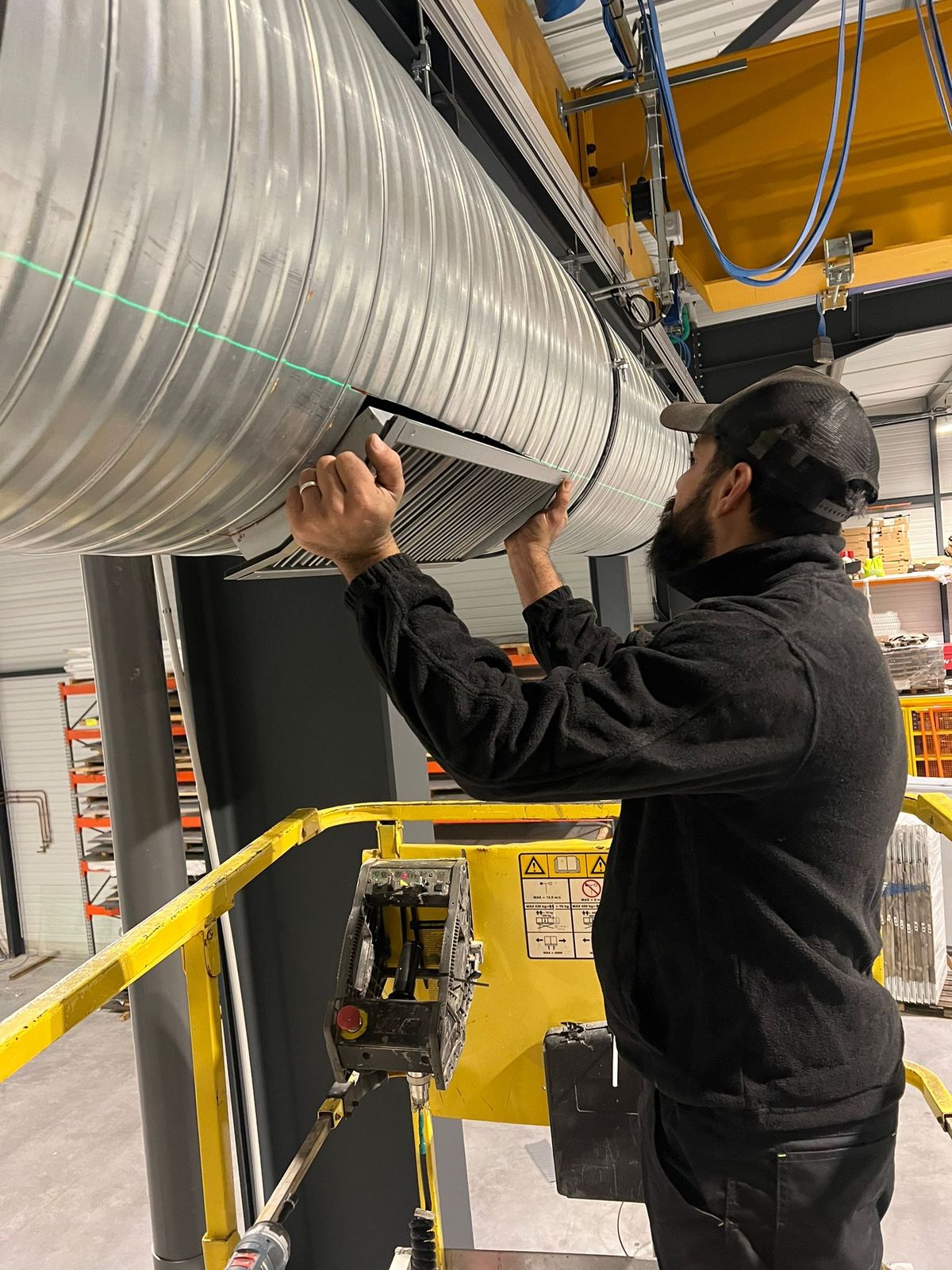 Technicien sur une nacelle, posant une grille sur une gaine spiralée dans un entrepôt